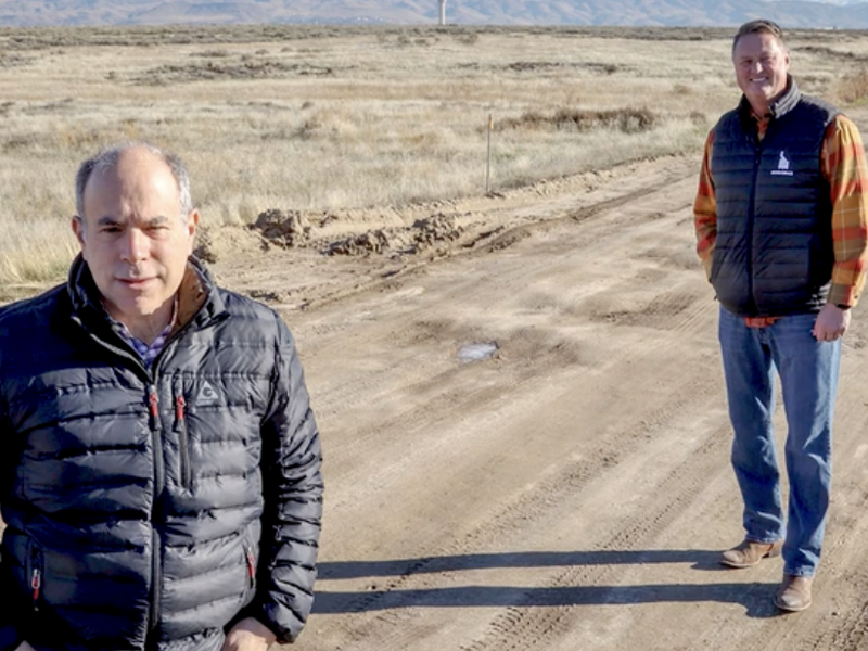 Two gentlemen in black vests on dirt road alongside dry field